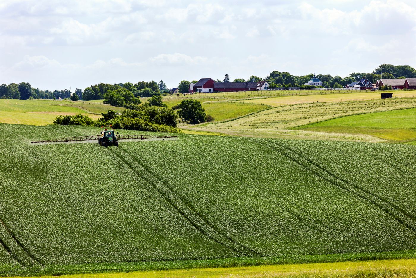 Miljöbild på en åker med en traktor. Bakom åkern står röda och vita hus