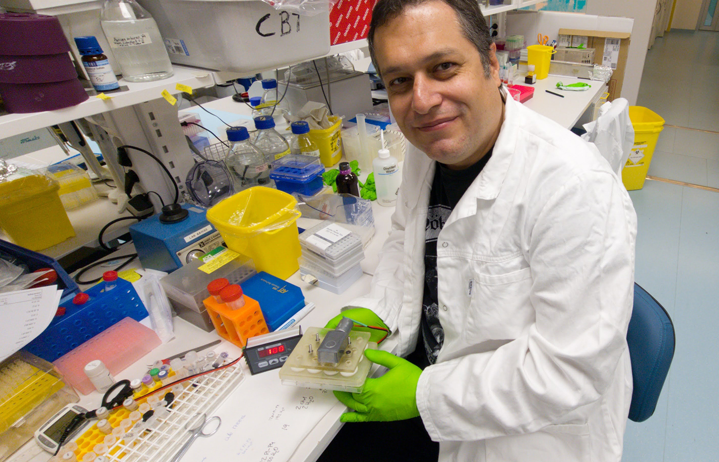 A man in the lab, dressed in lab protective clothes. He holds a plastic bowl for cell cultures. He looks straight into the camera.