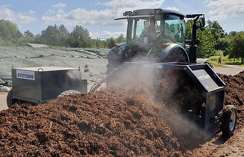 En mörk traktor och ett roterande redskap som bearbetar den avlånga högen av brunfärgad och ångande kompost.